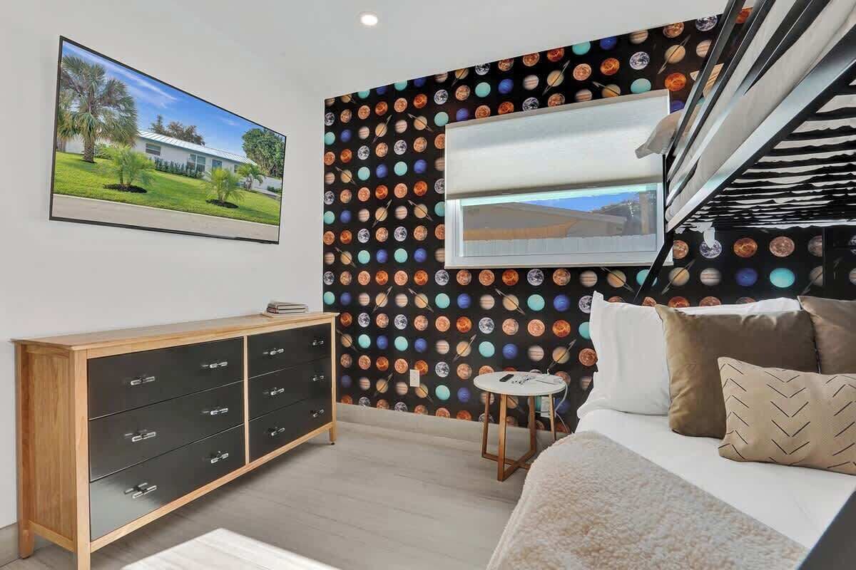 A bedroom with a bed, a dresser, a small table, and a wall with a space-themed wallpaper.