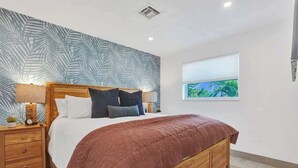 A bedroom with a wooden bed, a nightstand, a window with a view of greenery, and a patterned wall. - 5 mins to beach + Brand New Pool and Tiki Hut. (Oakland Park)