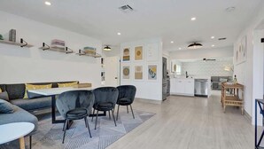 A modern living room with a sofa, chairs, a dining table, and a kitchen in the background. - 5 mins to beach + Brand New Pool and Tiki Hut. (Oakland Park)