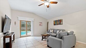 Un salon spacieux avec un canapé gris, un téléviseur posé sur un meuble en bois, un ventilateur de plafond et une porte-fenêtre coulissante donnant sur une piscine.