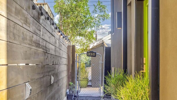 Une ruelle étroite bordée d’une clôture en bois, une porte métallique portant le numéro « 4451 », et une petite plante en pot.