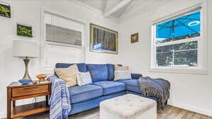 Un salon avec un canapé d’angle bleu, un pouf blanc, une table d’appoint en bois et une lampe. Il y a deux fenêtres équipées de stores, l’une donnant sur des arbres et l’autre sur un ciel bleu.