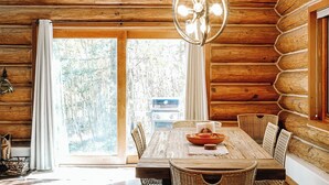 Interior de una cabaña de troncos con una mesa de comedor de madera, sillas de mimbre, una lámpara colgante y un ventanal con vistas a árboles.