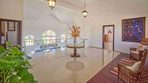 A spacious lobby with a high ceiling, a glass railing overlooking a lower level, a central round table with a vase of flowers, wicker furniture, and a large abstract painting on the wall. - Large Golf View Villa With Pool Jacuzzi and Staff (Punta Cana)
