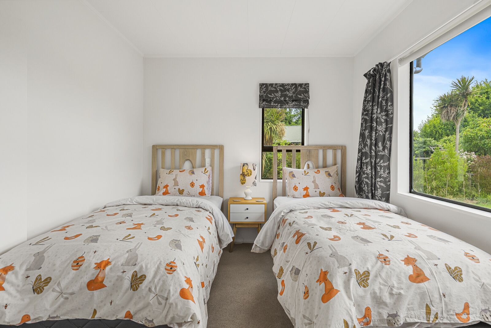 Quarto com duas camas, uma mesa de cabeceira, uma janela com vista para vegetação e uma janela com cortina estampada.