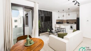 Een moderne woonkamer met een bank, een eettafel met een vaas, en een keuken met een koelkast en barstoelen.