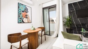 Een moderne woonkamer met een ronde houten tafel, twee houten stoelen, een bank, een groot abstract schilderij, een schuifdeur met toegang tot een balkon en een potplant.