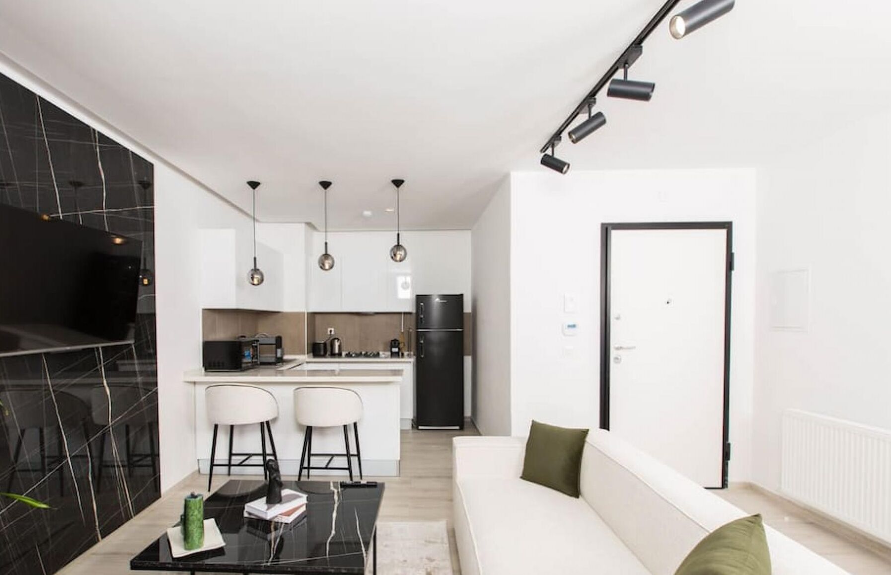 Una cocina moderna con un refrigerador negro, armarios blancos e iluminación colgante. Hay un comedor con una mesa negra y dos sillas blancas. En la sala de estar, hay un sofá blanco con cojines verdes.