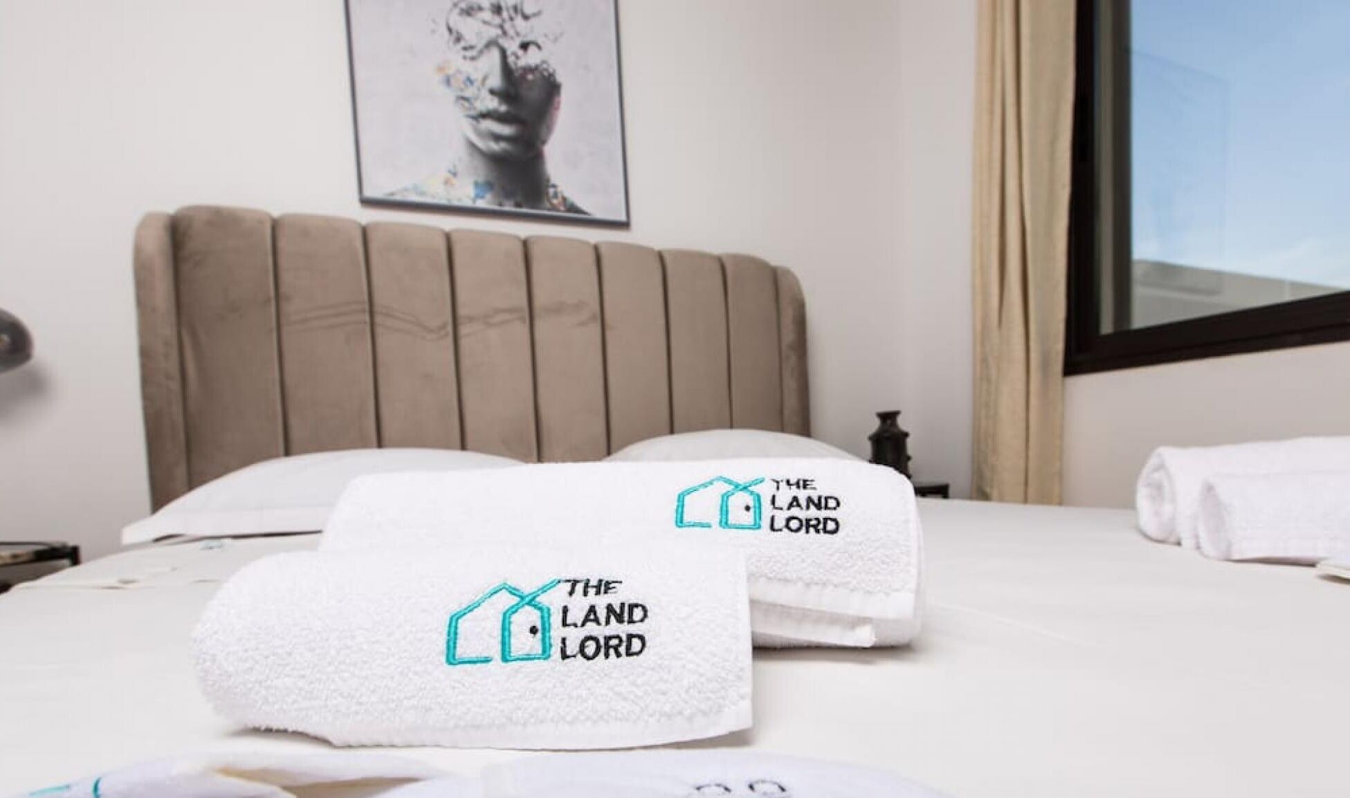 Una habitación de hotel con cama, dos toallas con el logotipo 'The Land Lord', cabecera de madera, una obra de arte enmarcada en la pared y una ventana con cortinas.