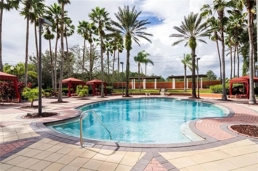 A swimming pool surrounded by palm trees and covered areas, with a tiled walkway and a clear sky.