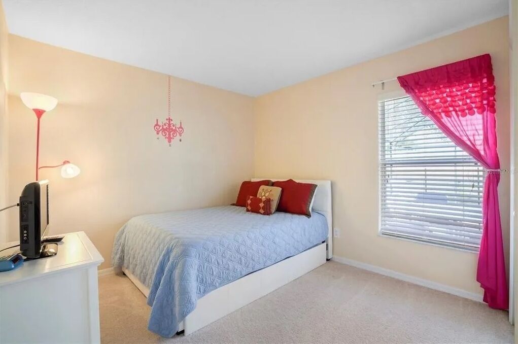 A bedroom with a bed, a desk with a computer, a lamp, and a window with blinds and curtains.