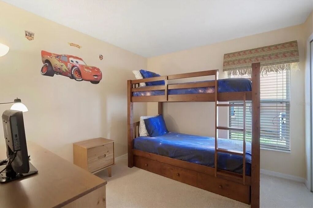 A bunk bed with a wooden frame and blue bedding, a desk with a lamp, a small drawer unit, and a wall decal of a race car.