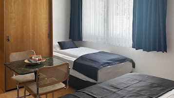 Una habitación compacta con una cama, una pequeña mesa con sillas, un armario de madera y una ventana con cortinas.