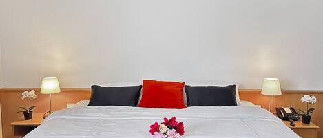A neatly made bed with a red pillow, white and black bedding, and a floral arrangement. There are two bedside tables with lamps and a telephone.
