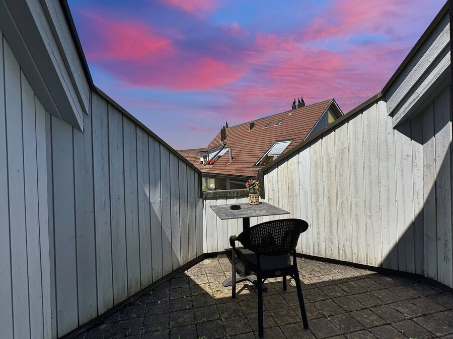 A covered patio with a table and chair, a view of the sky, and a building in the background.