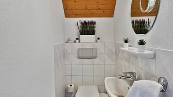 A modern bathroom with a wooden ceiling, white tiles, a toilet, a sink, a mirror, and small potted plants.