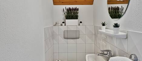 A modern bathroom with a wooden ceiling, white tiles, a toilet, a sink, a mirror, and small potted plants.