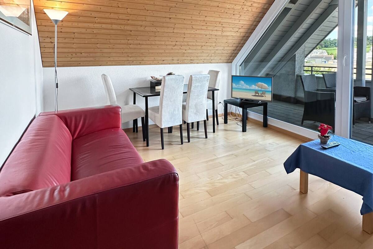 Ein modernes Loft mit Holzbalkendecke, einem roten Sofa, einem Esstisch mit Stühlen und Blick ins Freie durch große Fenster.