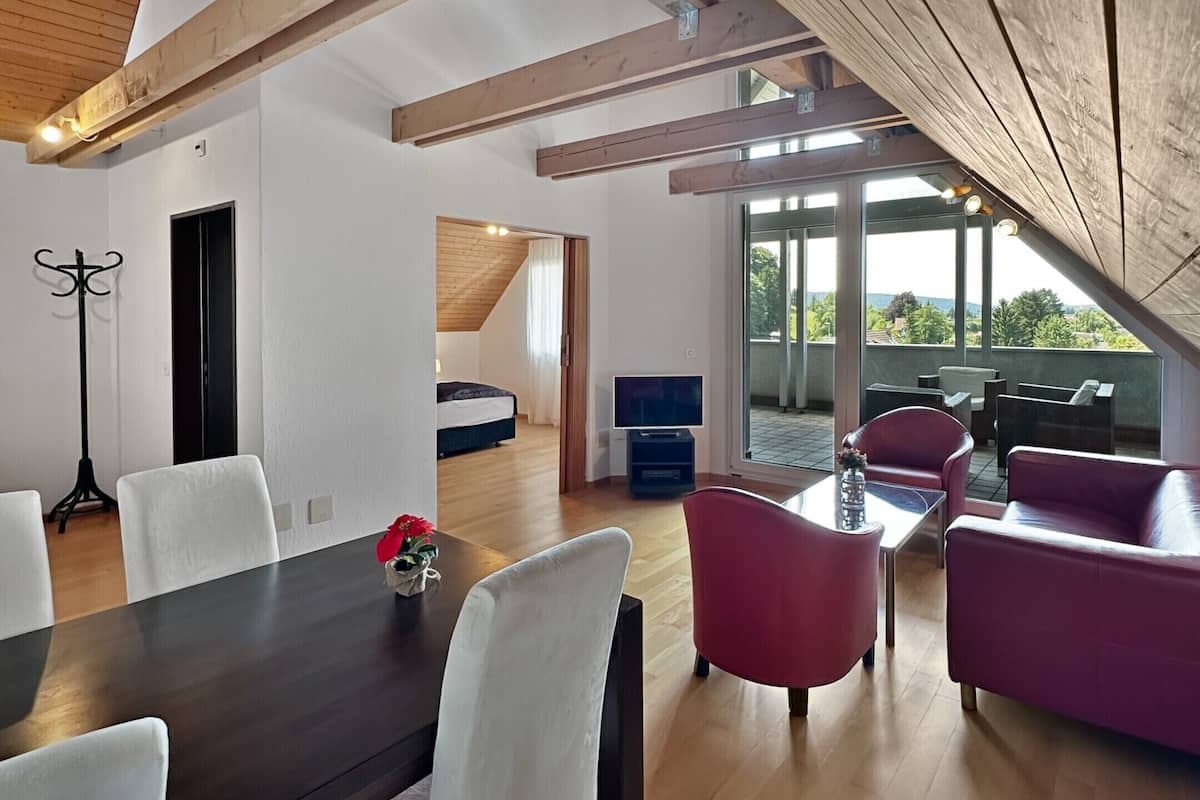 Ein modernes Wohnzimmer mit einem dunkelholznen Esstisch, weißen Stühlen und roten Sofas. Ein großes Fenster bietet einen Blick ins Freie.