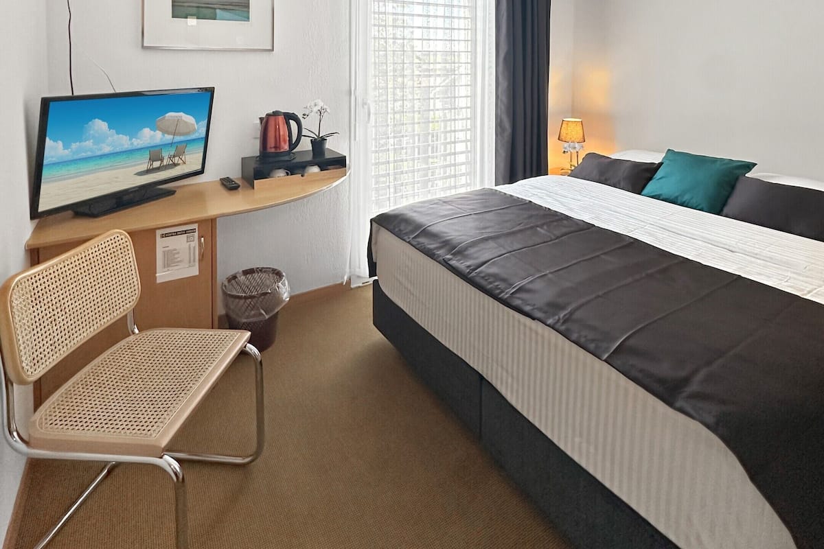 Ein Hotelzimmer mit einem Bett, einem Schreibtisch mit Computer, einem Sessel, einem Fernseher und einem Fenster mit Vorhängen.