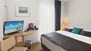 A hotel room with a bed, a desk with a computer, a chair, a TV, and a window with curtains.