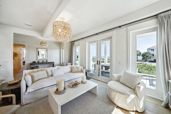 A spacious living room with a large sofa, a coffee table, and a chandelier. There is a balcony with a view of the ocean and a dining area with a table and chairs.