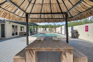 Un área junto a la piscina con una mesa de madera, sillas y una estructura de techo de paja.