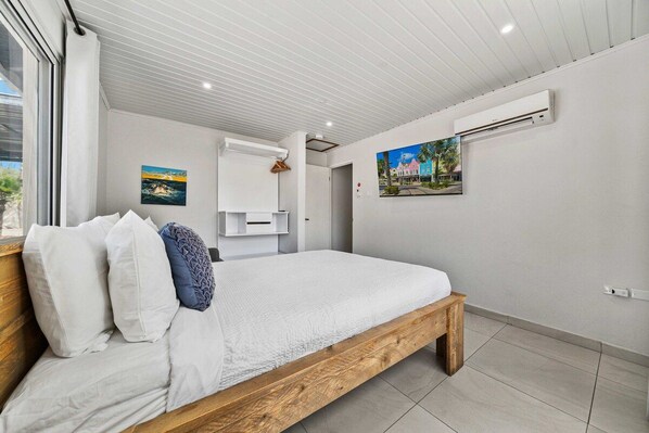 Un dormitorio con una cama de madera, ropa de cama blanca, una ventana y un equipo de aire acondicionado.