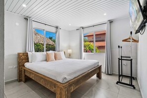 Ein Schlafzimmer mit einem hölzernen Bett, einem an der Wand montierten Fernseher und einem Fenster mit Blick auf ein Haus und Bäume.