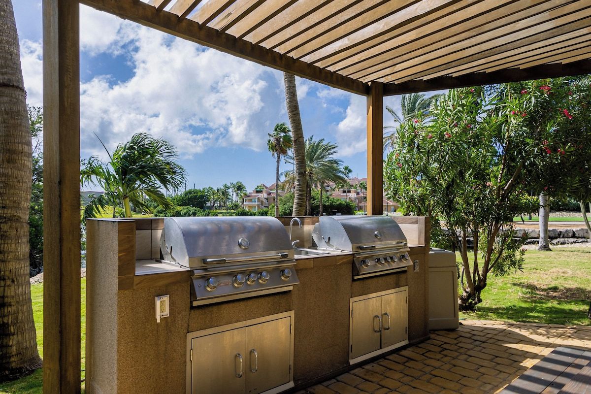 Un espace extérieur équipé d’un barbecue avec deux postes de cuisson, d’un coin salon abrité et d’où l’on peut admirer un complexe résidentiel en arrière-plan.