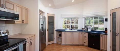 Una cocina moderna con armarios de madera clara, encimera negra, electrodomésticos de acero inoxidable y un ventanal que proporciona luz natural.