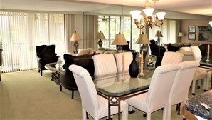 A hotel room with a dining area featuring a glass table surrounded by white upholstered chairs, a dark leather sofa, and a large mirror reflecting the room.