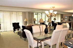 A hotel room with a dining area featuring a glass table surrounded by white upholstered chairs, a dark leather sofa, and a large mirror reflecting the room. - Gulf and Bay Club - C108 (Siesta Key)