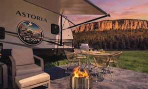 Un camper camperizzato con marchio 'ASTORIA THE DOVE', una zona pranzo con sedie e tavolo, e un braciere.