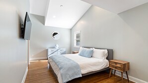 Une chambre avec un plafond en pente, un lit avec une tête de lit grise, une table de chevet avec un tiroir, un petit miroir rond et un téléviseur fixé au mur.