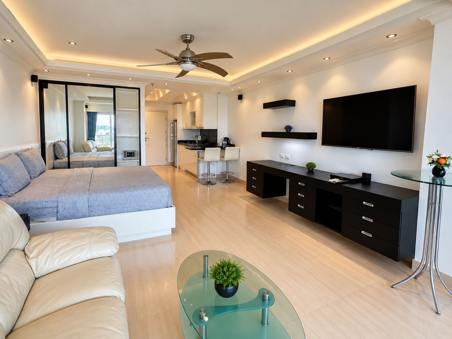 A modern bedroom with a bed, a glass coffee table, a TV, and a kitchen area in the background.