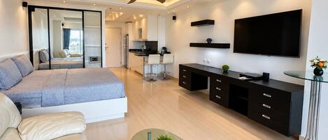 A modern bedroom with a bed, a glass coffee table, a TV, and a kitchen area in the background.