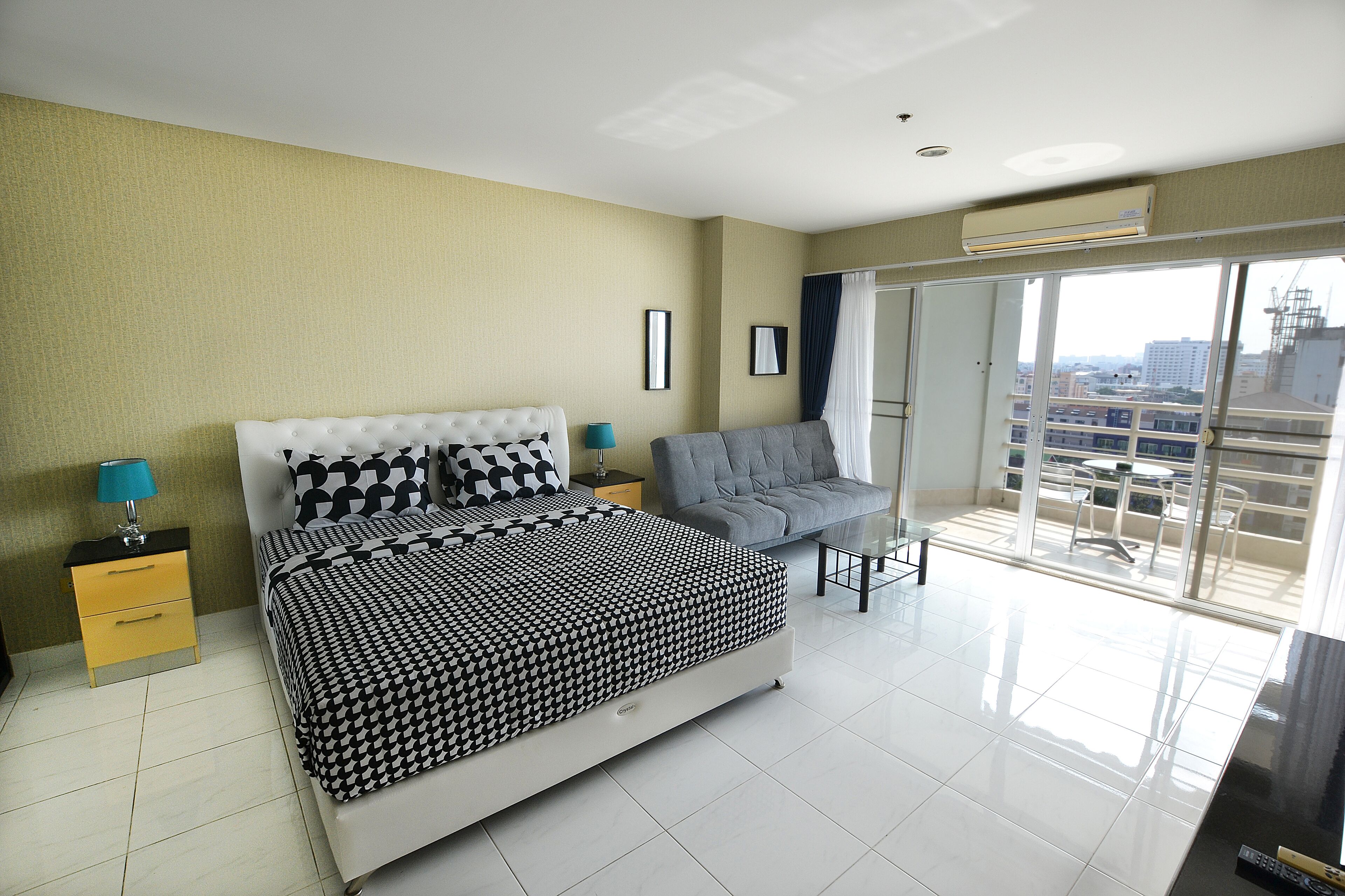 A modern bedroom with a large bed, a grey armchair, and a balcony with a city view.