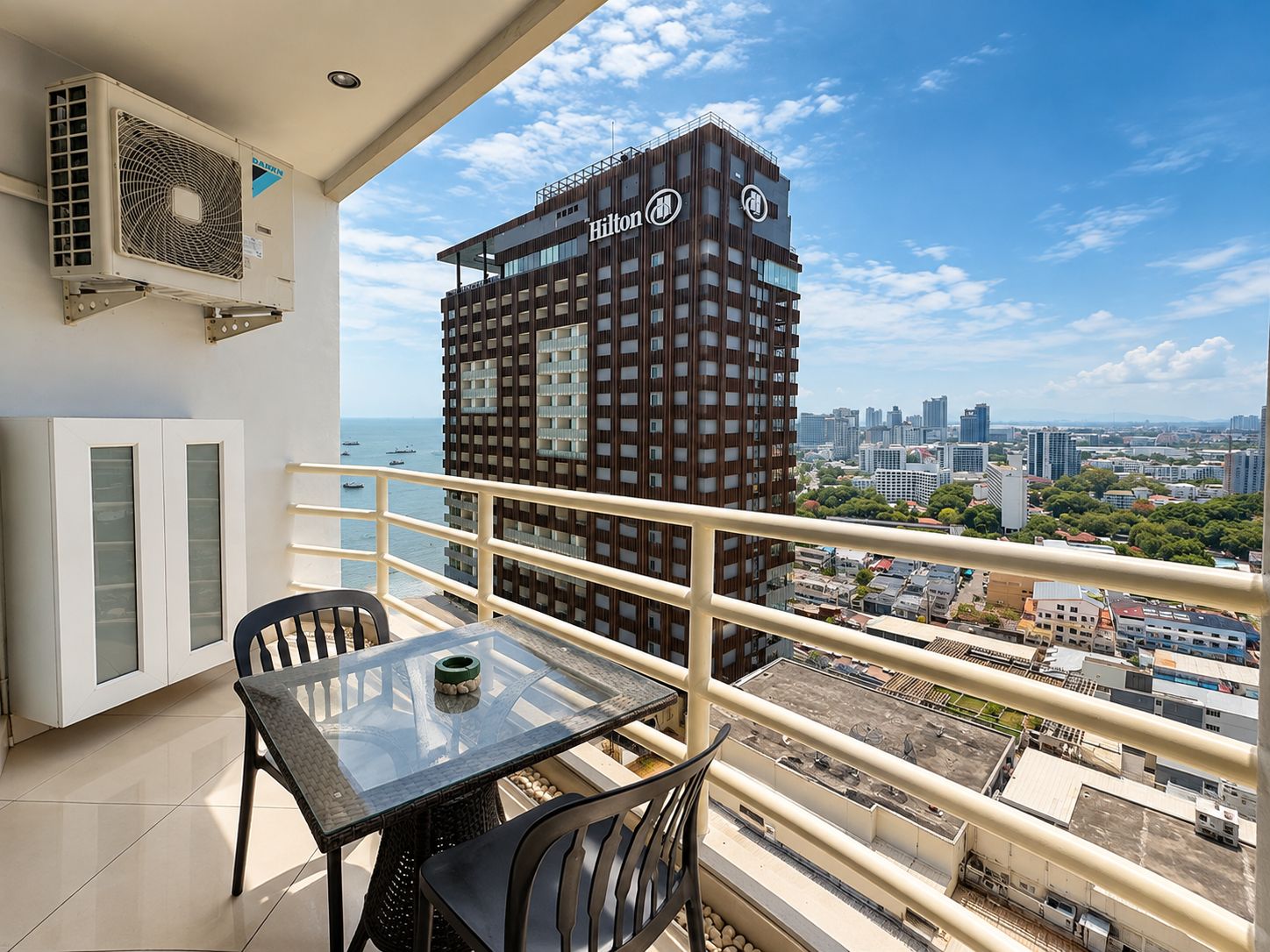 A balcony with a glass table and chairs, overlooking a cityscape with a prominent Hilton building.