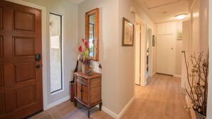 Un pasillo con una puerta de madera, una consola de mimbre con un espejo y un jarrón de flores.