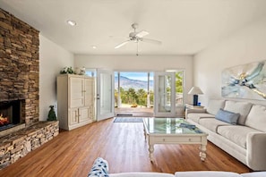 Un salon doté d’une cheminée en pierre, d’un canapé blanc, d’une table basse en verre et offrant une vue sur une terrasse extérieure, accessible par une porte vitrée.