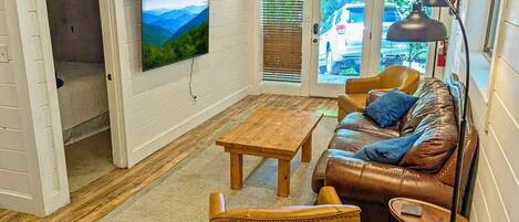 A living room with a wooden floor, a brown leather sofa, a wooden coffee table, and a television mounted on the wall.