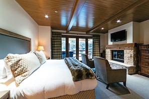 A bedroom with a large bed, a stone fireplace, a television, and a view of the outdoors.