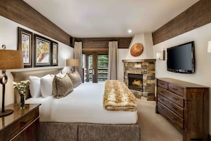 A bedroom with a large bed, a stone fireplace, a flat-screen TV, and framed artwork on the wall.