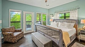 A bedroom with a bed, a chair, a bench, a ceiling fan, and a view of greenery through the windows.