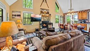 A spacious living room with a fireplace, large windows, and a dining area with a kitchen visible.