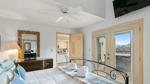 Ein Schlafzimmer mit Bett, Nachttisch, Lampe, Spiegel, Deckenventilator, Fernseher und Blick durch eine Tür.