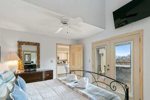 A bedroom with a bed, bedside table, lamp, mirror, ceiling fan, TV, and a view through a door.