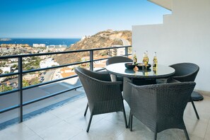 A balcony with a round table and four wicker chairs, overlooking a coastal cityscape.