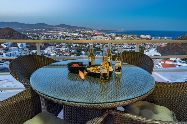 A round glass table with wicker chairs on a balcony overlooking a cityscape with mountains and the sea.